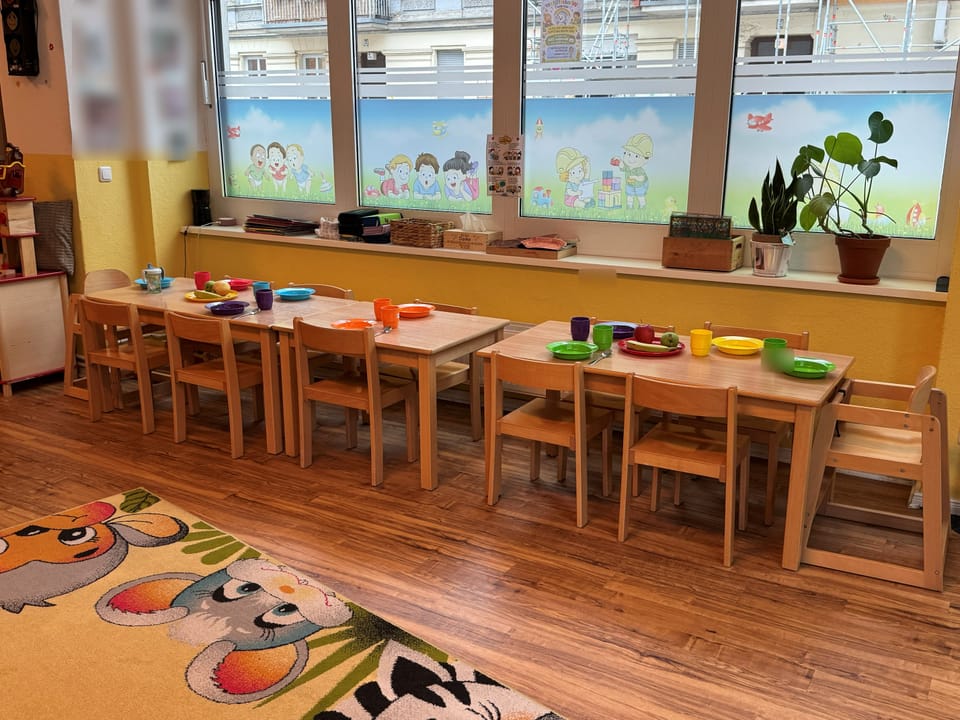 Langer, gedeckter Kindertisch mit bunten Bechern und Tellern vor großen Fenstern mit Kinder-Motiven