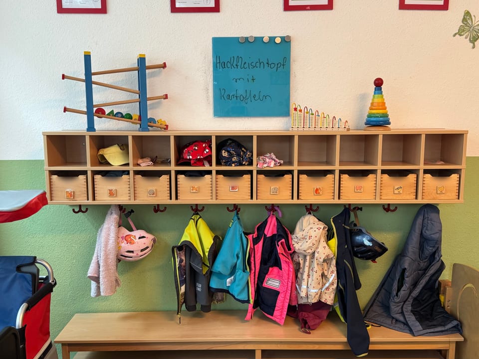 Garderobe mit Holzfächern, Jacken, Helmen und Schuhen der Kinder, auf der Tafel steht das Mittagessen