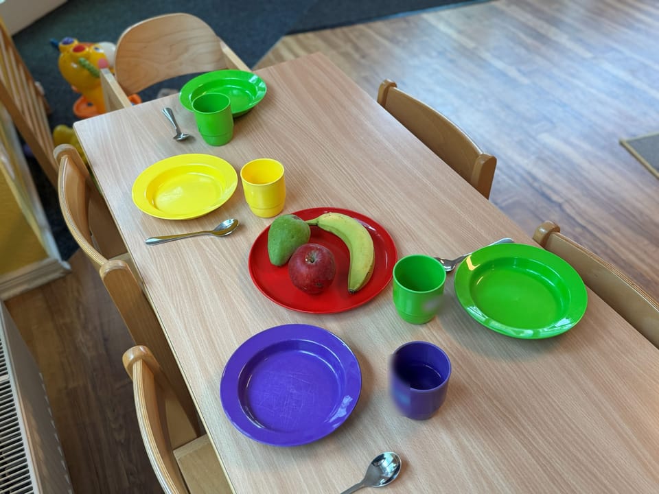 Bunte Teller und Becher, auf einem roten Teller liegen Apfel, Birne und Banane