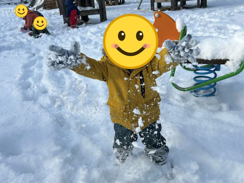 Ein Kind in gelber Jacke streckt im Schnee die Arme aus, im Hintergrund spielen weitere Kinder